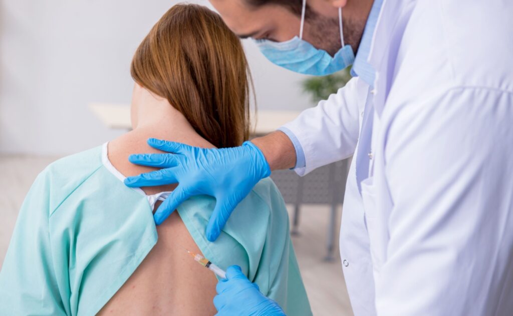 Ein Arzt injiziert einer Patientin ein Lokalanästhetikum in den Nackenbereich während einer Neuraltherapie-Sitzung zur Schmerzlinderung. Die Methode basiert auf der Theorie, dass gezielte Injektionen das autonome Nervensystem beeinflussen und Schmerzen lindern können.