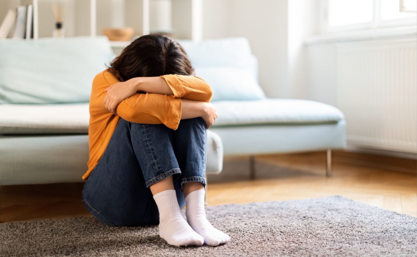 Junge Frau sitzt deprimiert auf dem Boden in einem hellen Wohnzimmer. Der Artikel beschreibt, wie Probiotika als ergänzende Therapie zu Antidepressiva bei der Behandlung von Depressionen helfen können.