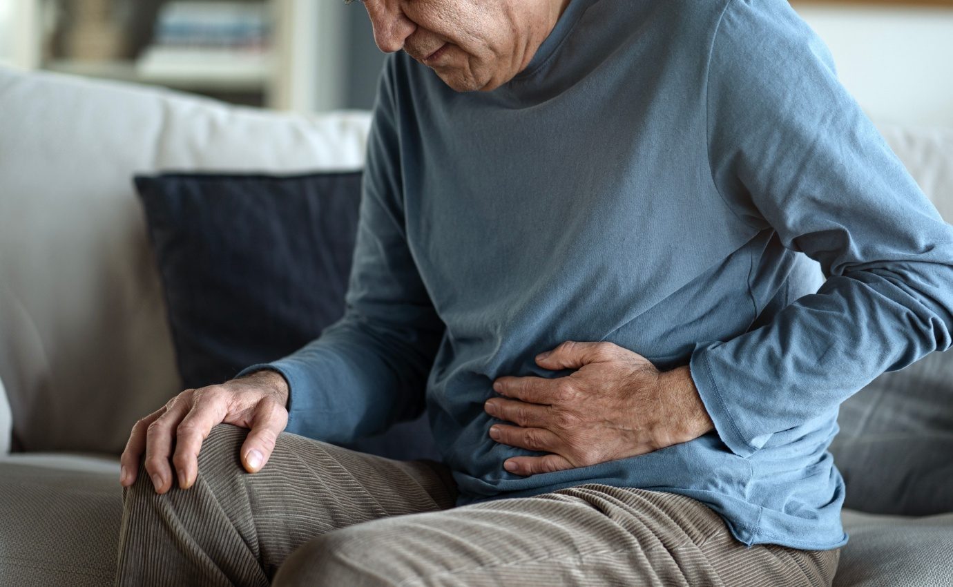 Ein älterer Mann sitzt auf einem Sofa und hält sich den Bauch vor Schmerzen. Der Artikel behandelt die potenziellen therapeutischen Vorteile von Myrrhe bei der Behandlung chronischer Darmerkrankungen wie dem Reizdarmsyndrom und Colitis ulcerosa.