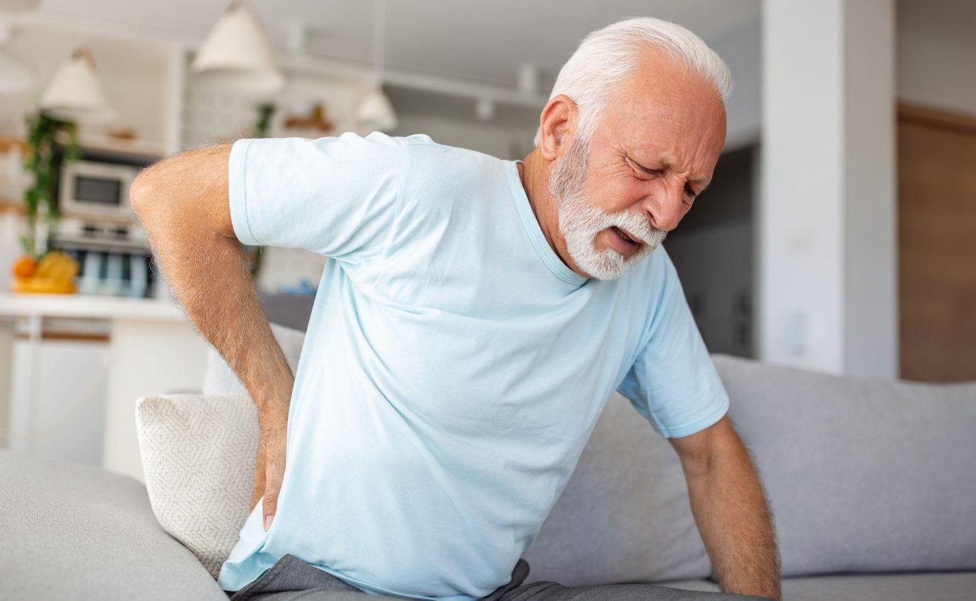 Ein älterer Mann sitzt auf einer Couch und hält sich schmerzverzerrt den unteren Rücken, was die Belastung durch chronische Schmerzen verdeutlicht. Der Artikel "Schmerzfrei ohne Chemie" beleuchtet nicht-pharmakologische Alternativen zur Schmerzbewältigung, wie Akupunktur und Bewegungstherapien.