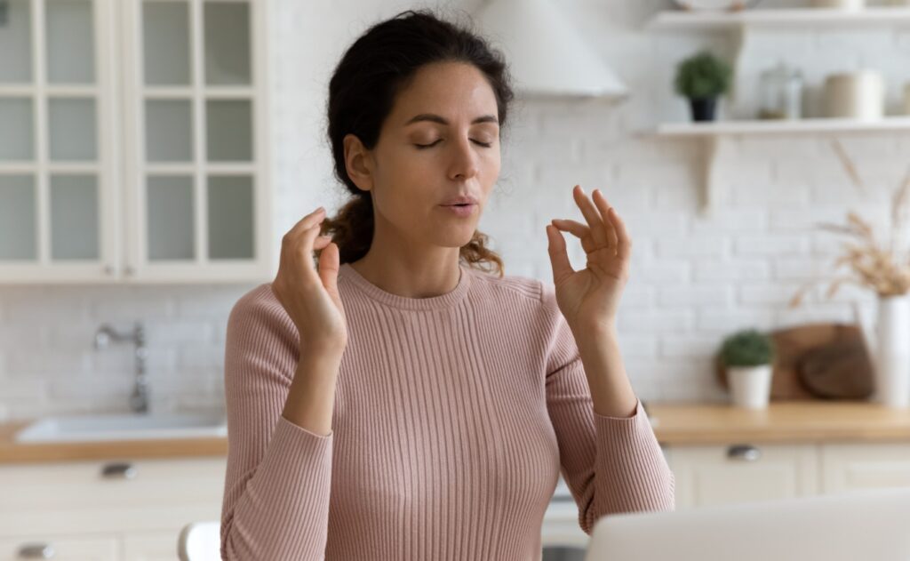 Eine Frau praktiziert eine Achtsamkeitsübung zur Stressbewältigung in ihrer Küche, um ihr Wohlbefinden zu verbessern. Der Artikel beleuchtet ganzheitliche und alternative Ansätze zur Reduktion von Stress.
