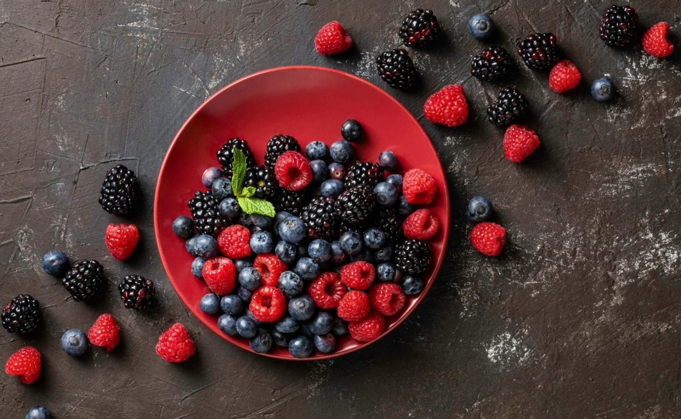 Eine bunte Auswahl an Beeren – Himbeeren, Brombeeren, Blaubeeren und Erdbeeren – auf einem dunkelroten Teller, arrangiert auf einem dunklen, strukturierten Hintergrund. Diese Lebensmittel sind reich an Flavonoiden, die laut einer aktuellen Studie in der Zeitschrift Nutrition & Diabetes das Risiko für Typ-2-Diabetes reduzieren können.
