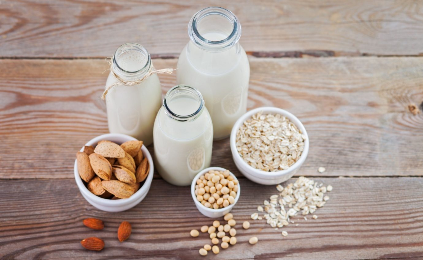Verschiedene Pflanzendrinks aus Hafer, Soja und Mandeln in Glasflaschen auf einem Holztisch. Diese pflanzlichen Milchalternativen bieten unterschiedliche Nährstoffprofile und sind eine beliebte Wahl für gesundheitsbewusste Menschen, die eine Alternative zu Kuhmilch suchen.