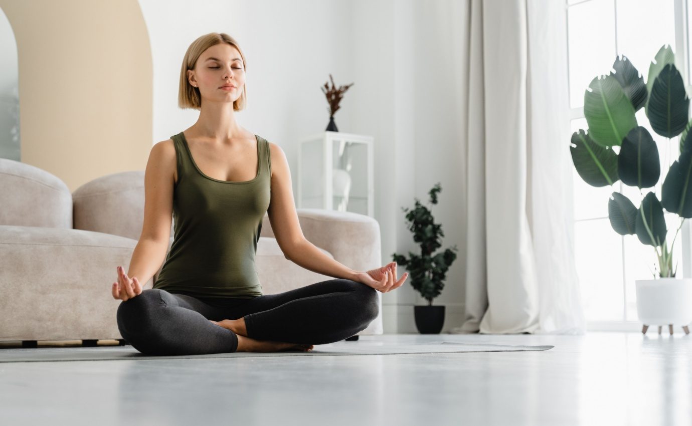 unge Frau in meditierender Haltung in einem hellen Wohnzimmer. Achtsamkeitsmeditation als wirksame Methode zur Schmerzreduktion und Stressbewältigung, unterstützt durch aktuelle Studien.