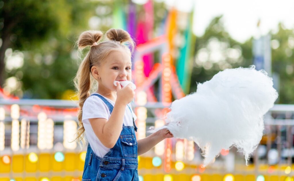 Kleines Mädchen isst Zuckerwatte auf einem Jahrmarkt, symbolisch für die Auswirkungen von Zuckerkonsum in der frühen Kindheit auf die langfristige Gesundheit.