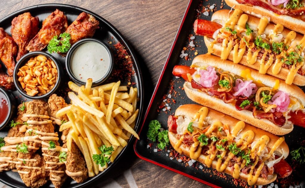 Nahaufnahme von hochverarbeiteten Lebensmitteln: Frittierte Hähnchenstreifen, Pommes, Chicken Wings, Hot Dogs mit Käse, Speck und eingelegten Zwiebeln. Die neue Studie zeigt, dass der übermäßige Konsum solcher Ultra-Processed Foods (UPFs) das Sterberisiko erhöhen kann.