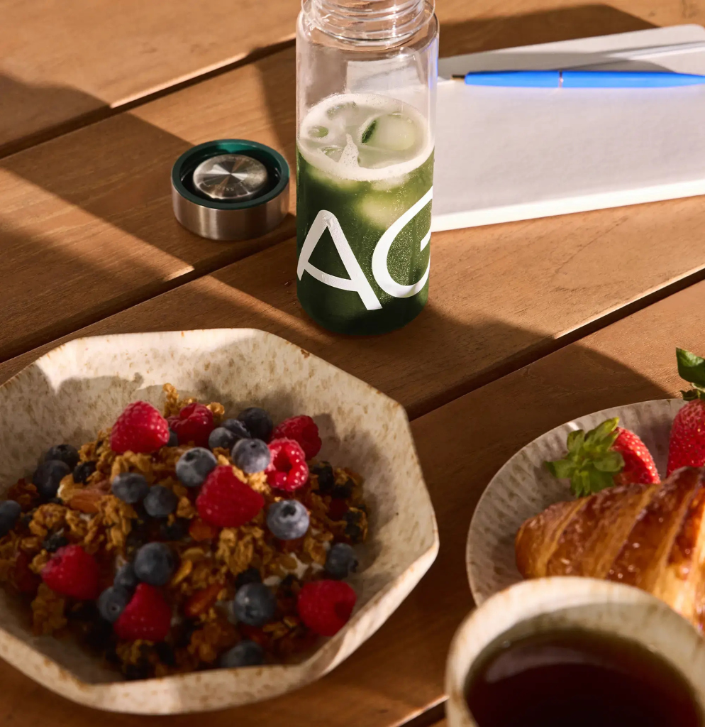 AG1-Flasche mit grünem Drink auf Frühstückstisch mit Beerenmüsli, Croissant und Tee