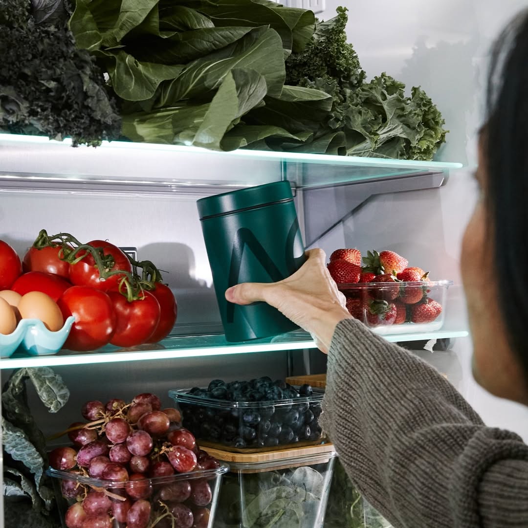 Hand stellt AG1-Dose in gut gefüllten Kühlschrank mit frischem Obst und Gemüse