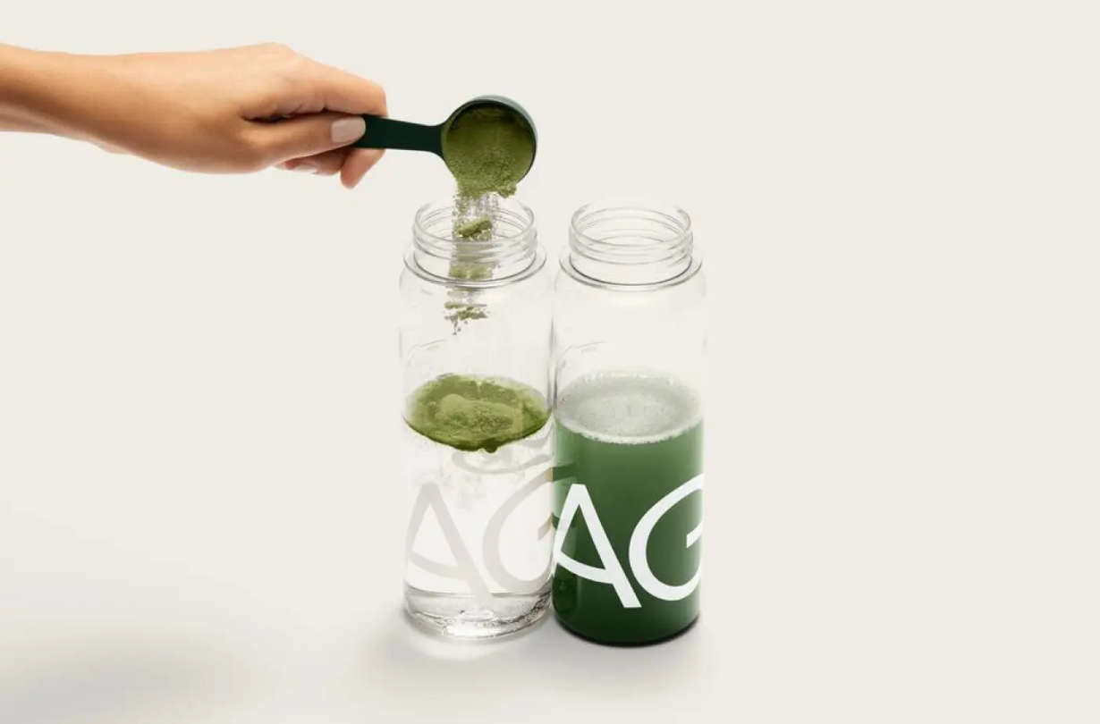 Hand gießt grünes AG1-Pulver in ein Glas Wasser – das beliebte Nahrungsergänzungsmittel mit über 70 Inhaltsstoffen für Energie, Verdauung und Immunsystem.