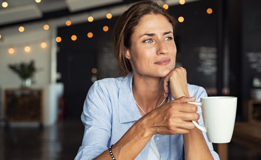 Eine nachdenklich wirkende Frau mittleren Alters sitzt mit einer Tasse Kaffee in einem Café – neue Studie zeigt, dass regelmäßiger Kaffeekonsum bei Frauen das gesunde Altern fördern kann.